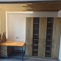 MAKKU Mateusz Kułak - Drewniana szafa z przeszkleniami i biurko z szufladami, industrialne nogi, dekoracyjna drewniana zabudowa sufitu i ściany, nowoczesne wnętrze.