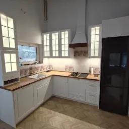 Jasna kuchnia na wymiar z drewnianym blatem i przeszklonymi szafkami wiszącymi, z czarną lodówką w zabudowie. Podłoga wyłożona płytkami imitującymi parkiet.