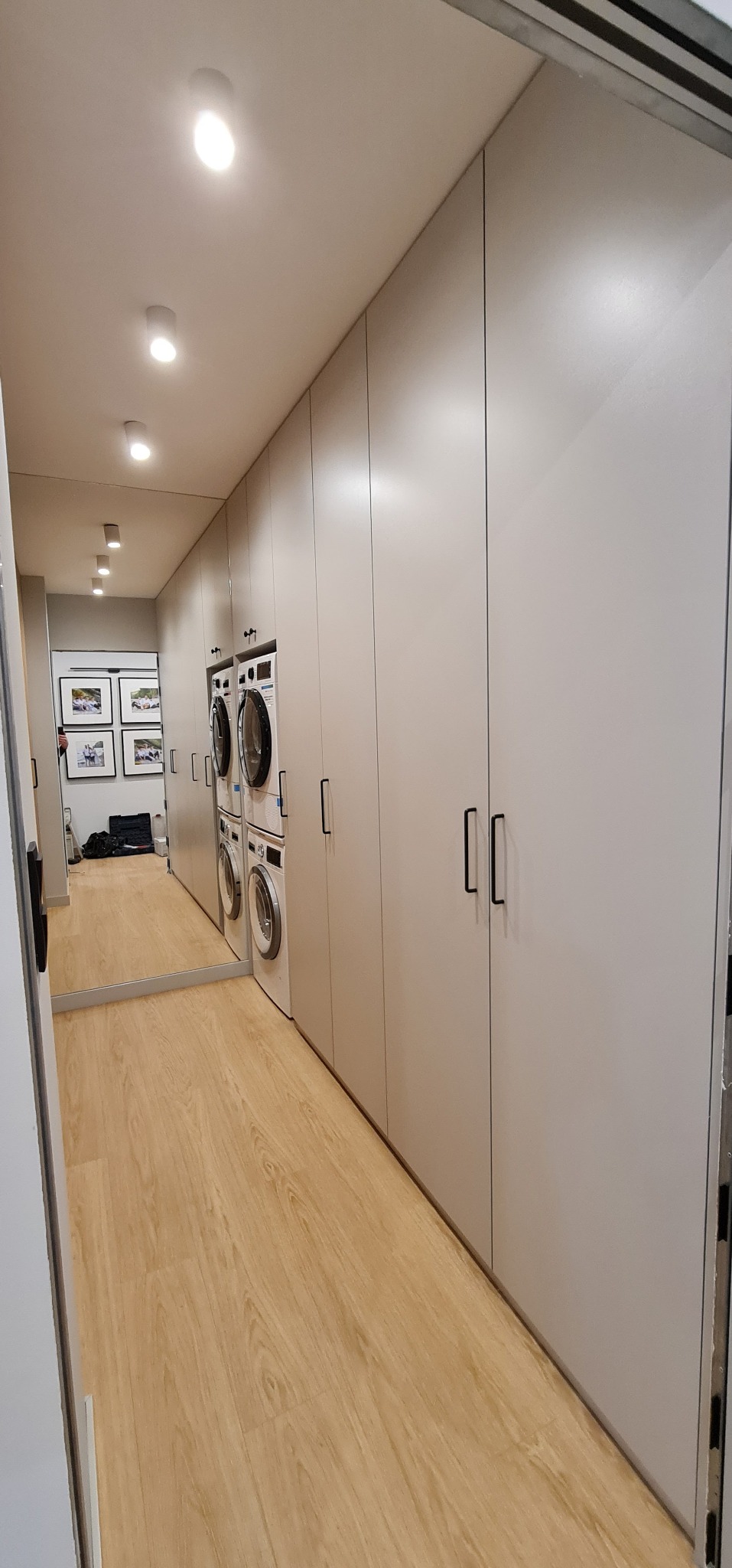 Długa, szara szafa wnękowa z czarnymi uchwytami, integrująca pralkę i suszarkę, z lustrem na końcu korytarza i drewnianą podłogą.