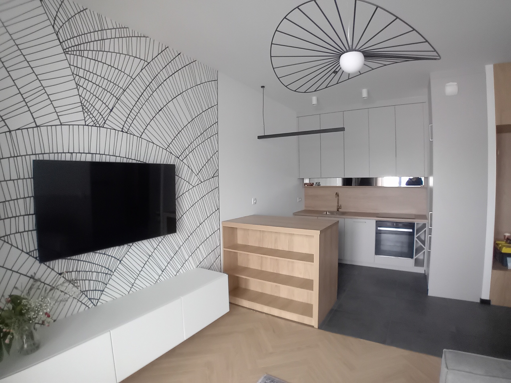 Nowoczesna kuchnia z wyspą i geometryczną tapetą. Minimalistyczne szafki, designerskie lampy i drewniane akcenty tworzą spójną, stylową przestrzeń.