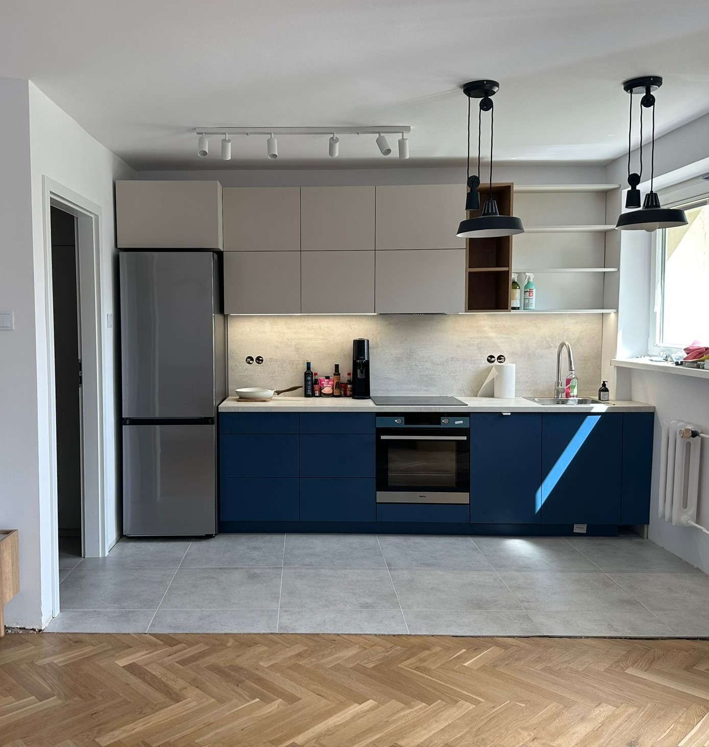 Nowoczesna kuchnia z granatowymi szafkami dolnymi i szarymi górnymi, oświetlona lampami wiszącymi i listwą LED, na tle parkietu i szarych płytek podłogowych.