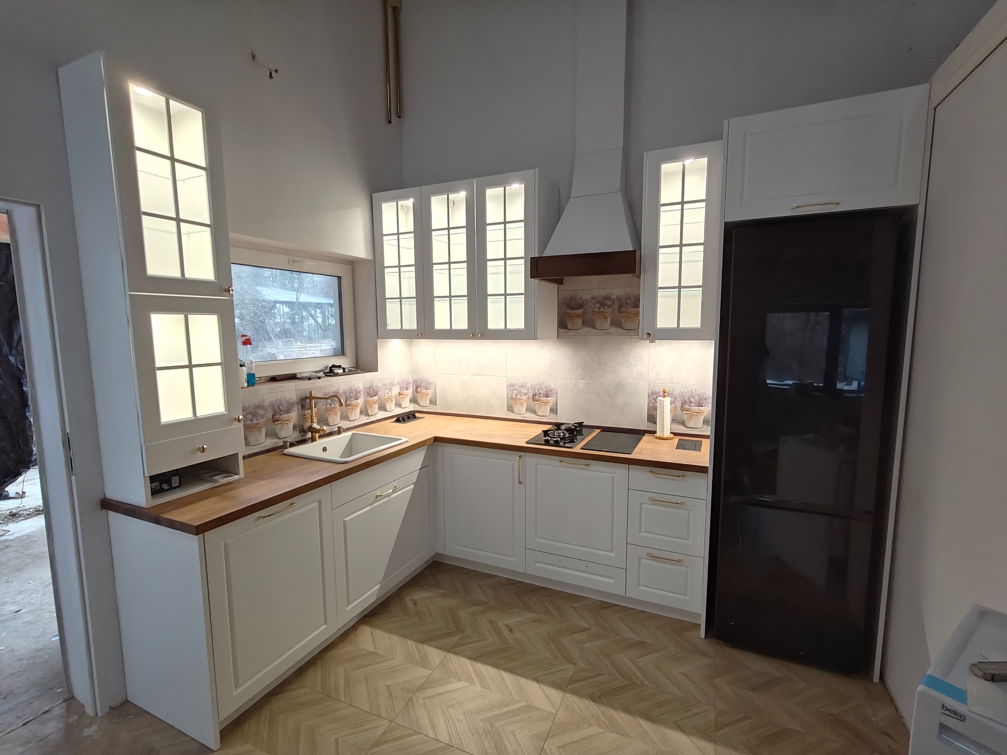 Jasna kuchnia na wymiar z drewnianym blatem i przeszklonymi szafkami wiszącymi, z czarną lodówką w zabudowie. Podłoga wyłożona płytkami imitującymi parkiet.