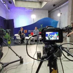 Ekipa filmowa kręci wywiad z dwoma mężczyznami w studio z niebieskim tłem i logiem miasta Łódź, widok zza kamery z operatorem ustawiającym ostrość.