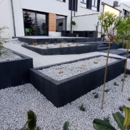 Nowoczesny ogród z podwyższonymi rabatami obłożonymi czarną kostką, wypełnionymi białym grysem i młodymi roślinami, z kostki brukowej wykonane ścieżki i schody, na tle białego domu z dużymi oknami.