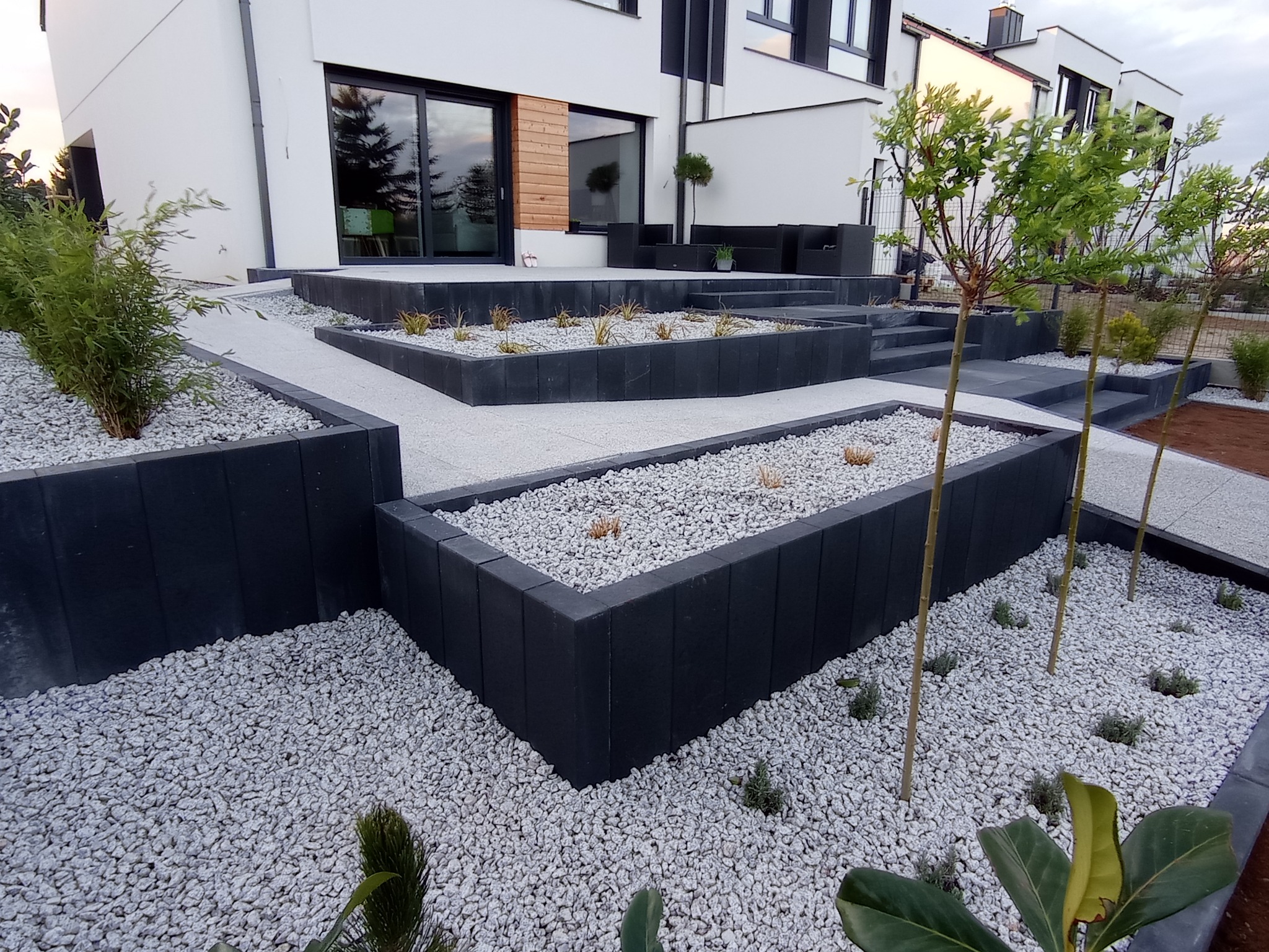Nowoczesny ogród z podwyższonymi rabatami obłożonymi czarną kostką, wypełnionymi białym grysem i młodymi roślinami, z kostki brukowej wykonane ścieżki i schody, na tle białego domu z dużymi oknami.