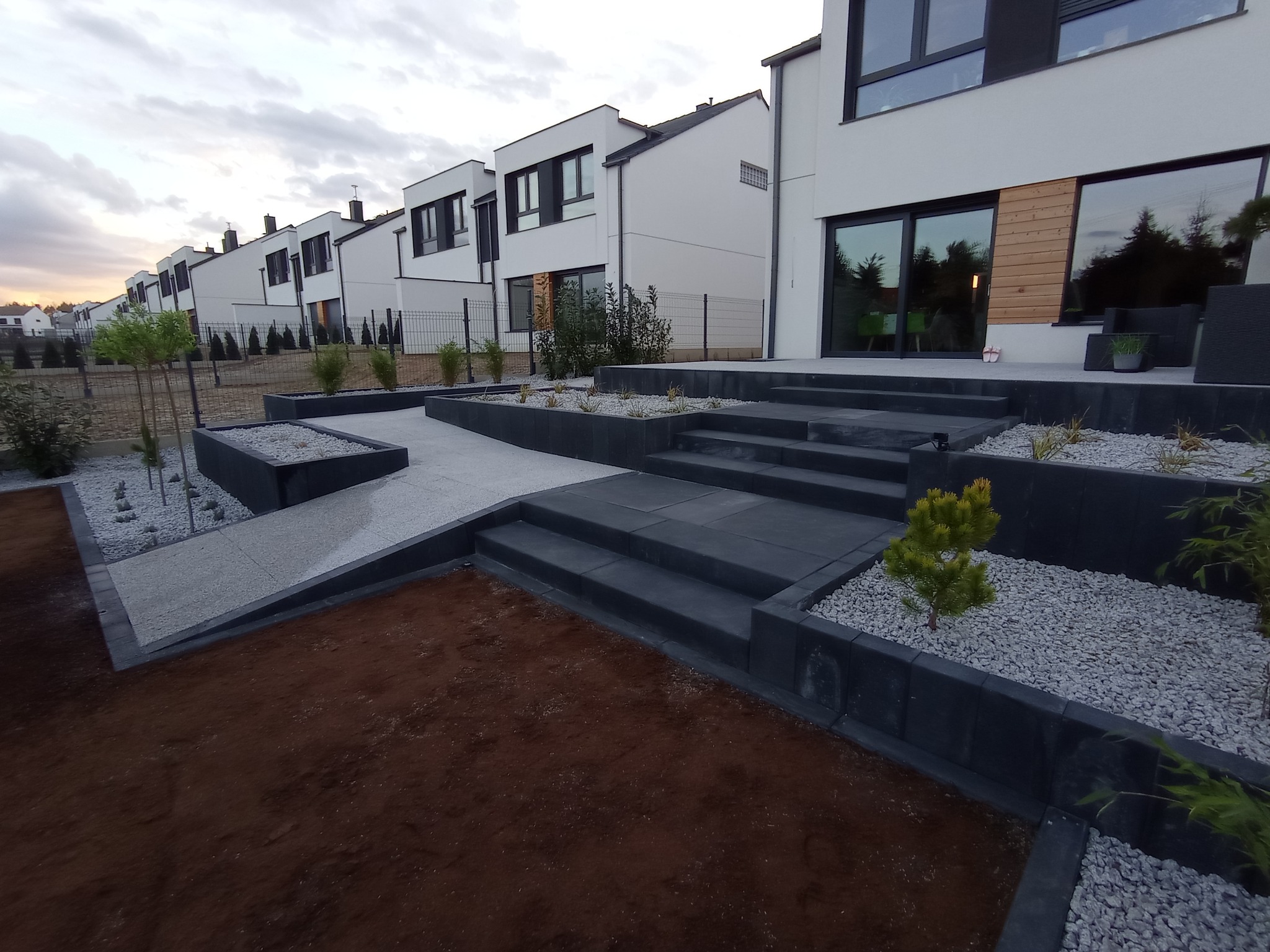 Nowoczesny ogród przed szeregiem domów jednorodzinnych: schody z ciemnej kostki, podwyższone rabaty z szarą żwirową podsypką i młodymi roślinami, świeżo przygotowana gleba pod trawnik.