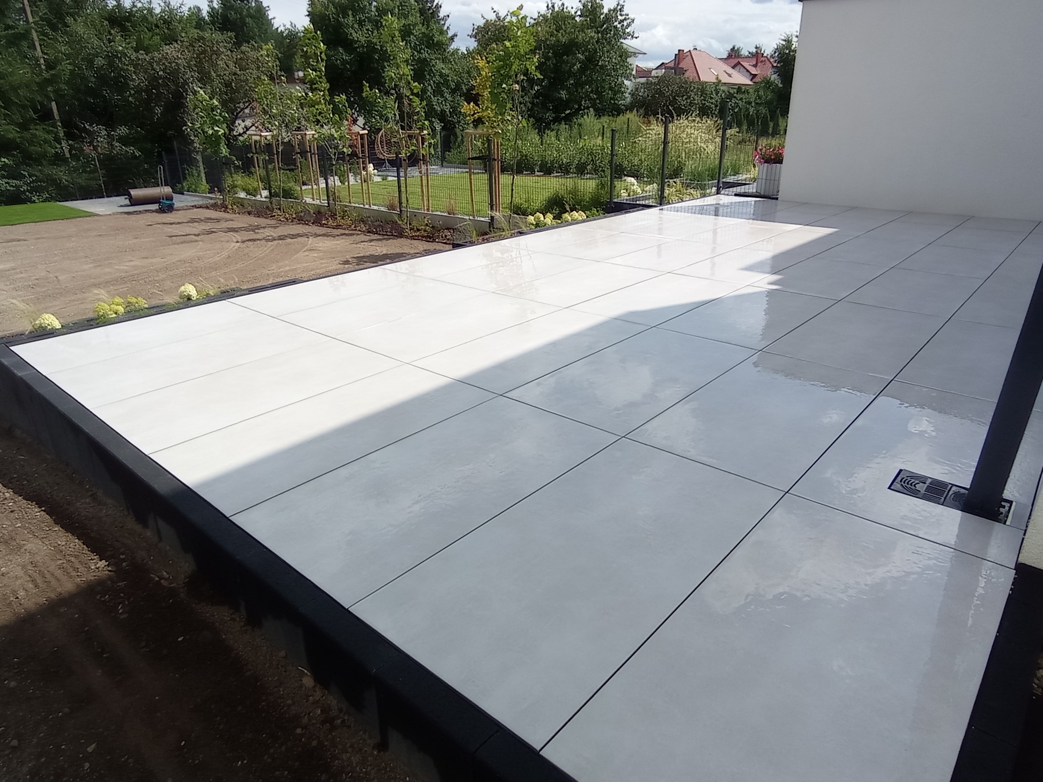 Świeżo ułożony, jasny taras z dużych płyt betonowych, z odpływem liniowym i widokiem na ogród z młodymi drzewkami.