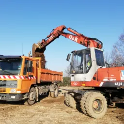 Pomarańczowa wywrotka Volvo FL10 pod koparką O&K, która wsypuje ziemię do skrzyni ładunkowej na placu budowy w słoneczny dzień.