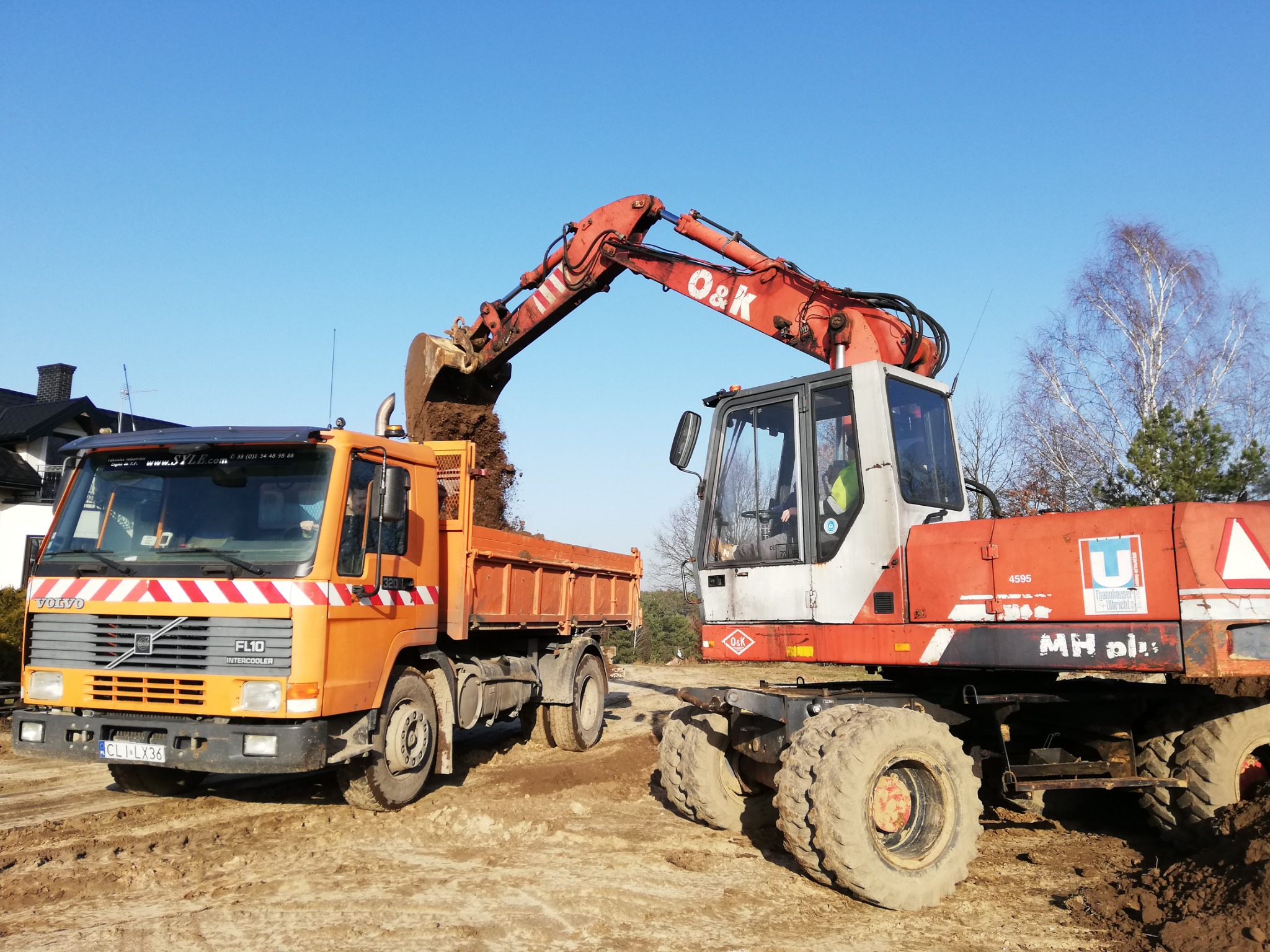 Pomarańczowa wywrotka Volvo FL10 pod koparką O&K, która wsypuje ziemię do skrzyni ładunkowej na placu budowy w słoneczny dzień.