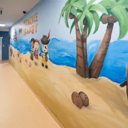 Korytarz z muralem przedstawiającym piracką tematykę: postacie piratów, palmy, morze i napis 'Pirackie skarby' w jasnych, pastelowych kolorach. Widoczne drzwi do innych pomieszczeń.