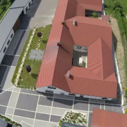 Widok z lotu ptaka na budynek szwalni z czerwonym dachem i wewnętrznym dziedzińcem, otoczony zielenią i parkingiem z geometrycznym wzorem kostki brukowej.