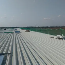 Widok z góry na rozległy, przemysłowy dach z blachy trapezowej w trakcie montażu, z widocznymi pracownikami w oddali i zabezpieczeniami siatkowymi na obrzeżach, w tle słupy wysokiego napięcia...