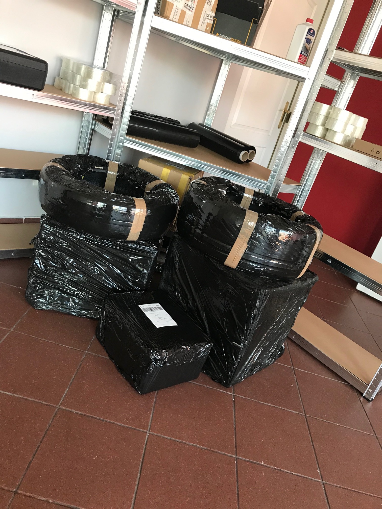 Paczki owinięte czarną folią i taśmą leżące na brązowej podłodze z płytek, z metalowym regałem w tle.