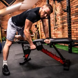 Mężczyzna w krótkich spodenkach i czarnej koszulce ćwiczy z hantlami na ławce w siłowni z ceglaną ścianą w tle.