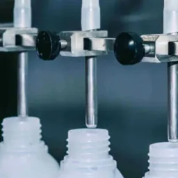 Automatyczne napełnianie plastikowych butelek z wąską szyjką, widoczne metalowe dysze i regulowane uchwyty.