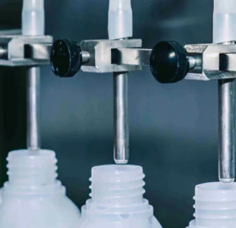 Automatyczne napełnianie plastikowych butelek z wąską szyjką, widoczne metalowe dysze i regulowane uchwyty.