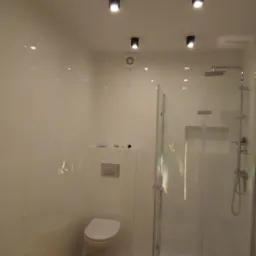 Nowoczesna łazienka z białymi panelami ściennymi, kabiną prysznicową ze szklanymi drzwiami, podwieszaną toaletą i minimalistycznym oświetleniem sufitowym.