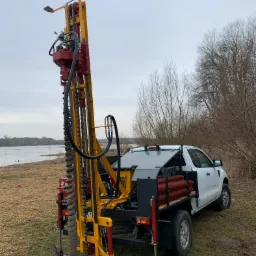 Wiertnica zamontowana na pick-upie podczas wiercenia w gruncie, widoczny krajobraz z jeziorem w tle.