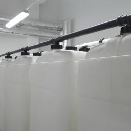 SKY WAY LEON WAWRENIUK - Rząd białych zbiorników z czarnymi pokrywami i rurami w jasnym pomieszczeniu technicznym z widocznymi rurami wentylacyjnymi pod sufitem.