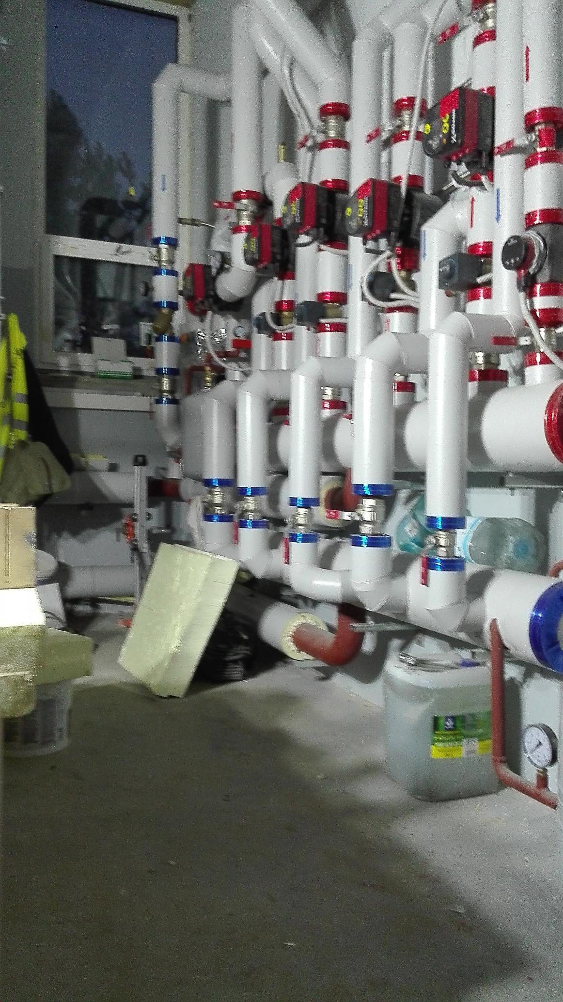 Złożona instalacja rur grzewczych z zaworami i czujnikami w pomieszczeniu technicznym, widoczne materiały izolacyjne i kanister z płynem.