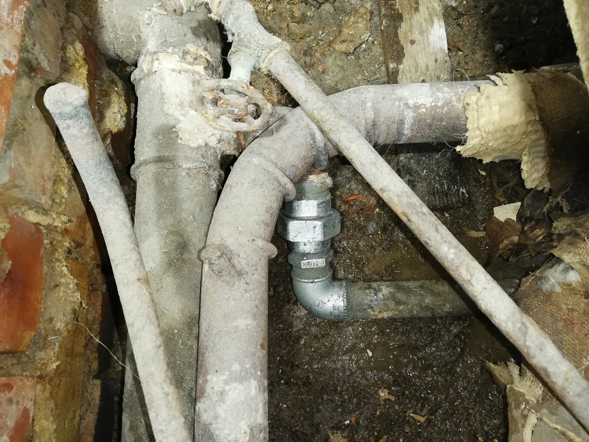 Zardzewiałe rury hydrauliczne z zaworem i nowym złączem w piwnicy, widoczny fragment ceglanej ściany i fragmenty izolacji