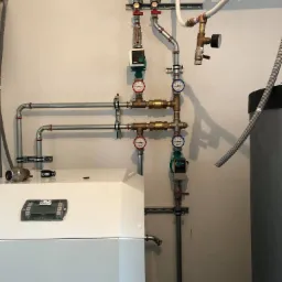 Kocioł grzewczy z widocznymi rurami, zaworami, manometrami i pompami obiegowymi, zamontowany na ścianie w pomieszczeniu technicznym.
