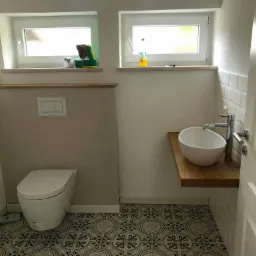 Nowoczesna łazienka z wiszącą miską WC, umywalką nablatową na drewnianym blacie i wzorzystą podłogą, oświetlona przez dwa okna.