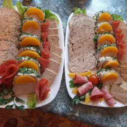 Dwa białe półmiski z pasztetem, salami, pieczywem i brzoskwiniami, udekorowane zieloną pietruszką i sałatą, na tle marmurowego blatu.