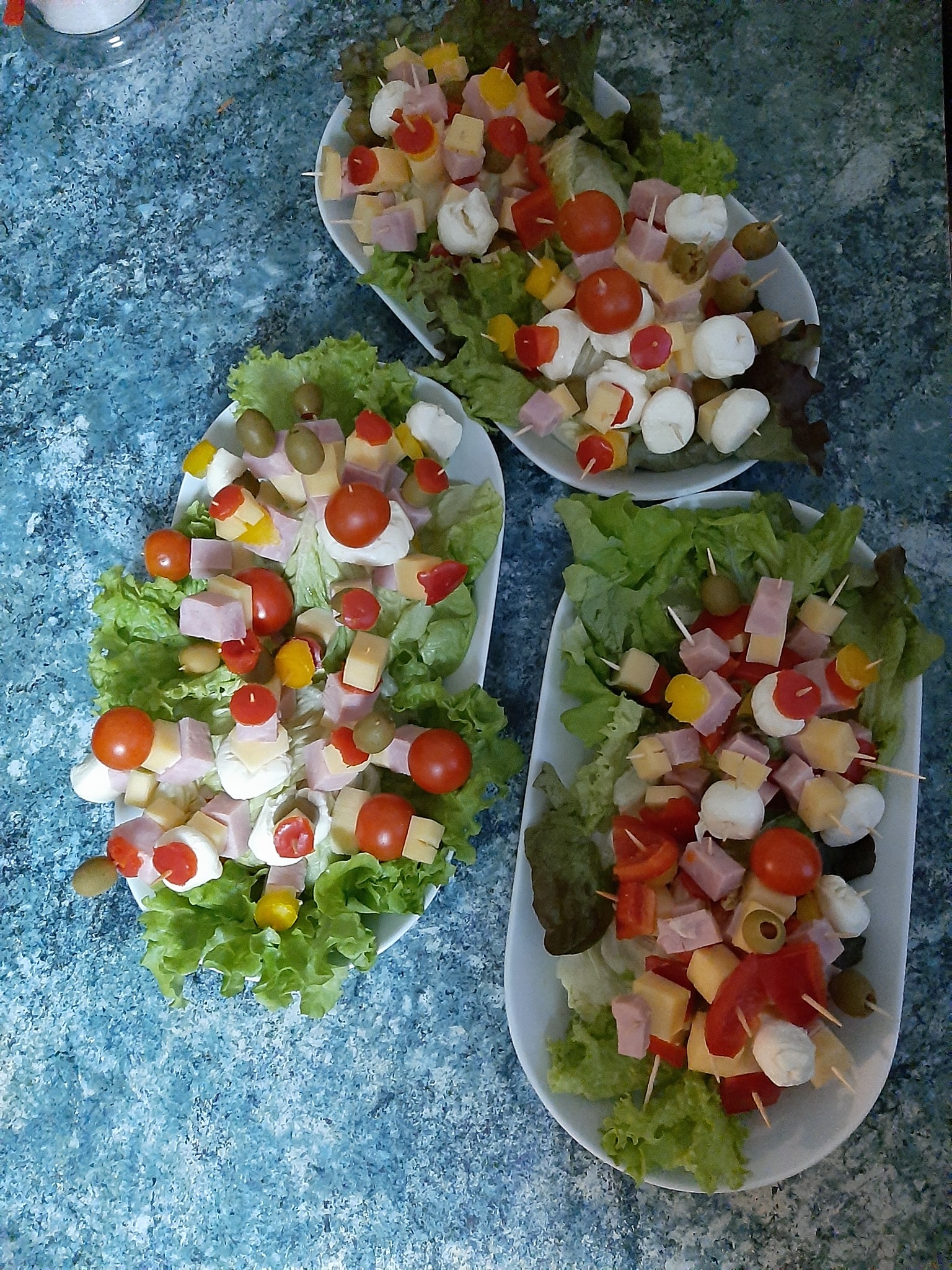 Trzy białe półmiski z przekąskami na szpilkach: kulki mozzarelli, pomidorki cherry, kostki szynki i sera, oliwki, papryka, ułożone na liściach sałaty.