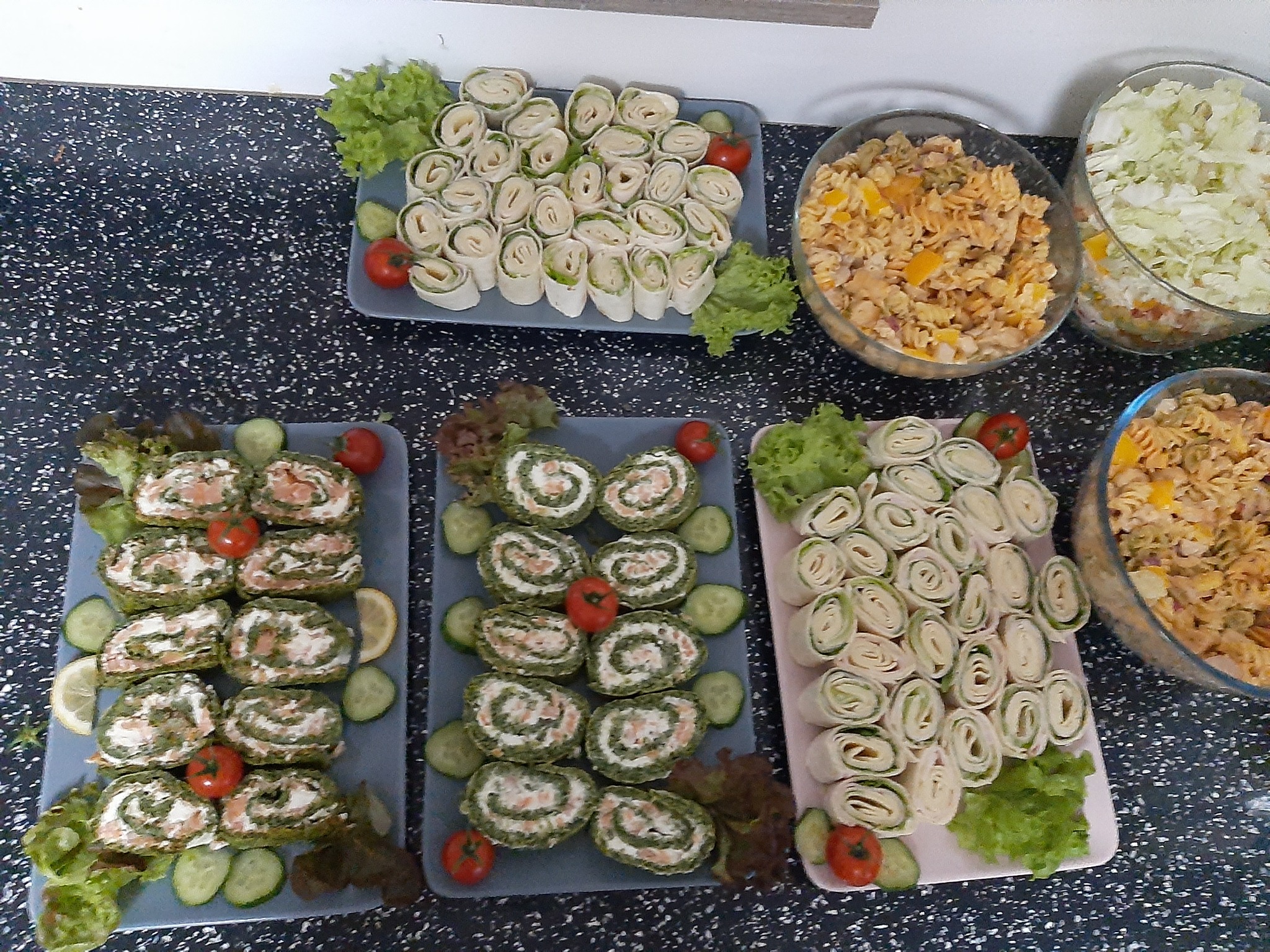 Różnorodne roladki i sałatki makaronowe ułożone na talerzach i w miskach, prezentacja cateringowa na czarnym blacie.