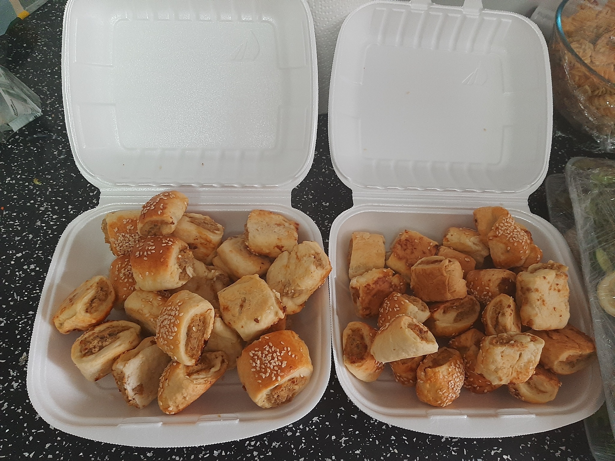 Dwa białe pojemniki styropianowe wypełnione roladkami z ciasta francuskiego i mięsnym farszem, posypane sezamem, widoczne na czarnym blacie.