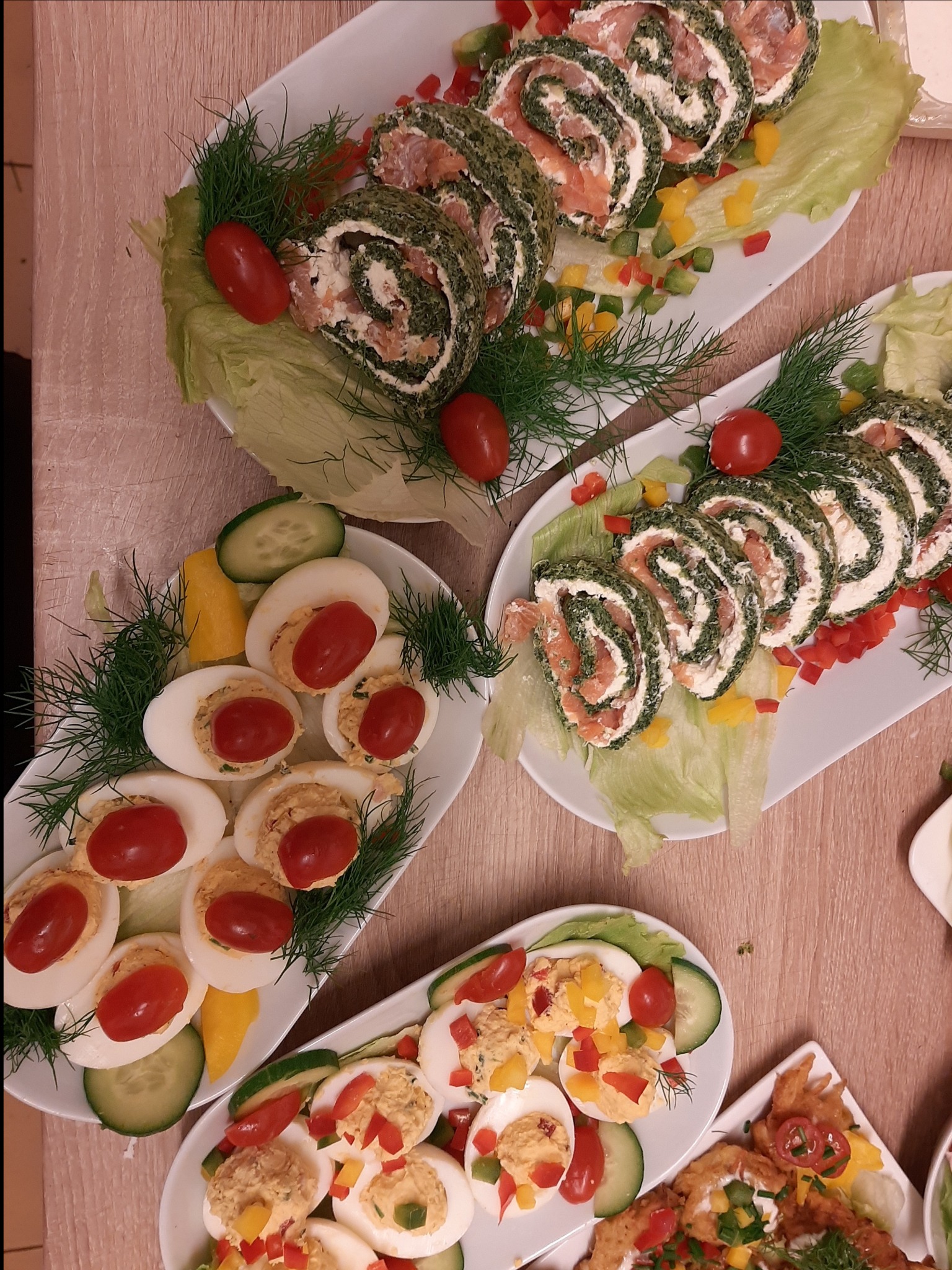 Dekoracyjnie ułożone na białych talerzach przystawki: roladki ze szpinakiem i łososiem, jajka faszerowane z pomidorkami koktajlowymi, udekorowane koperkiem i warzywami.