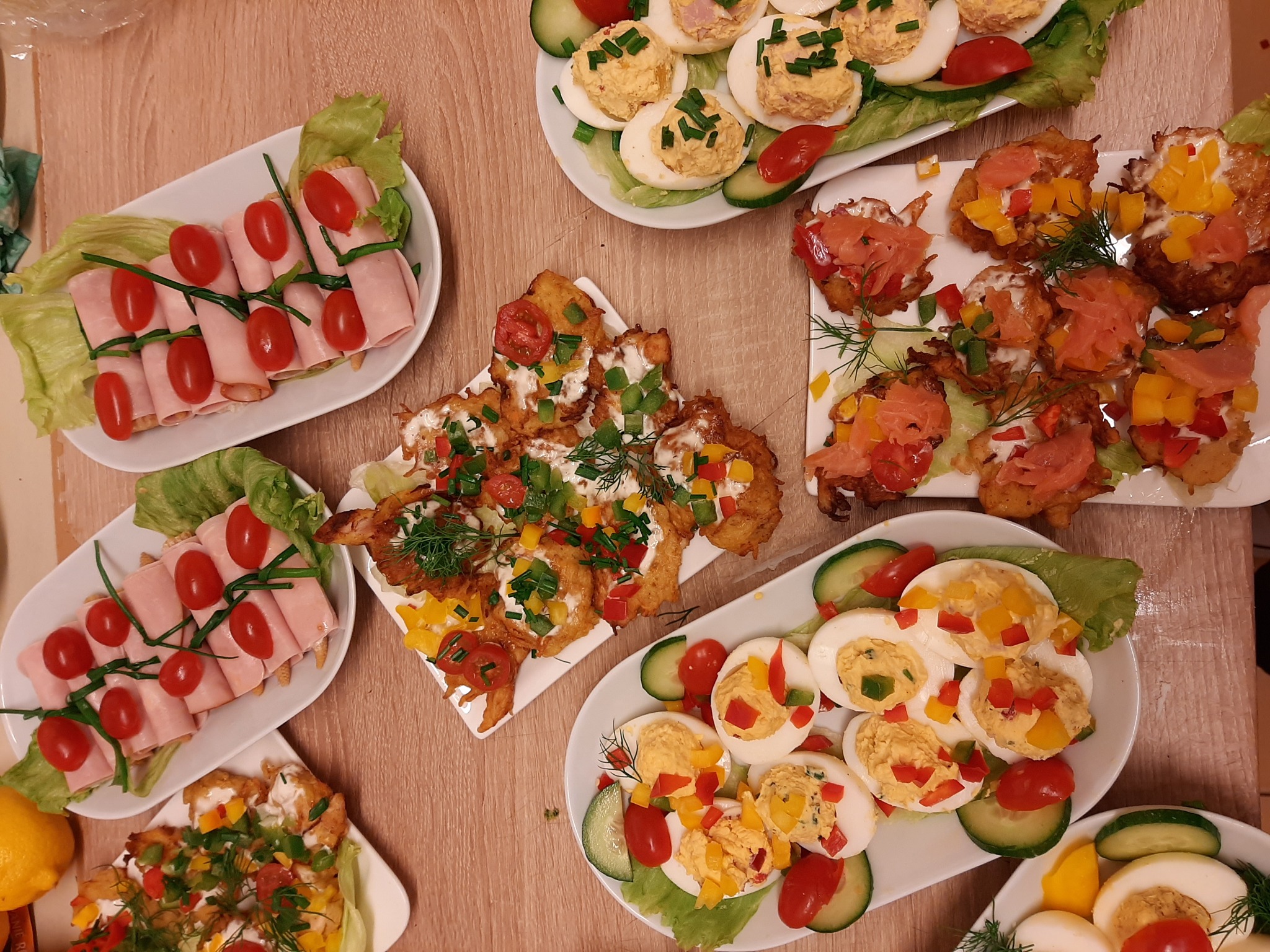 Dekoracyjnie ułożone przekąski na białych talerzach: roladki z szynki z pomidorkami cherry, jajka faszerowane z papryką, placki ziemniaczane z łososiem i sosem, wszystko udekorowane zieleniną...