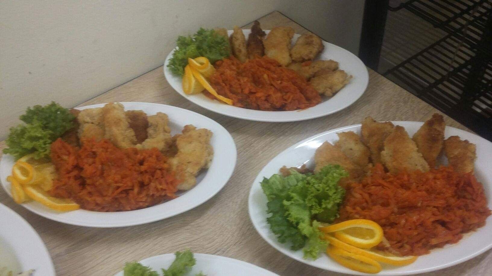 Trzy talerze z panierowaną rybą, surówką z marchwi i dekoracją z sałaty oraz pomarańczy, ustawione na stole.