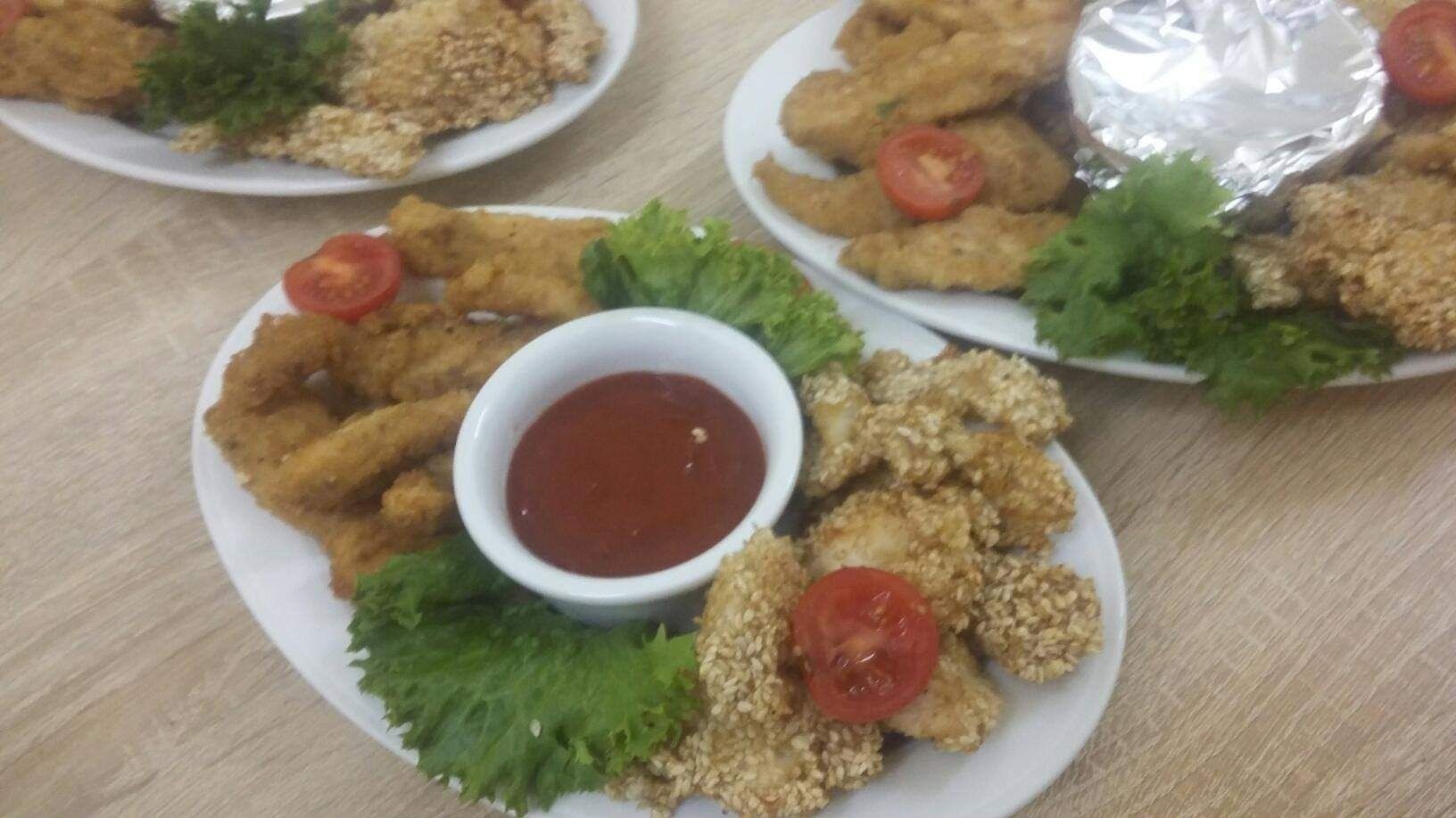 Trzy talerze z nuggetami z kurczaka, część z nich obtoczona w sezamie, podane z sosem pomidorowym, pomidorkami cherry i sałatą na drewnianym stole.