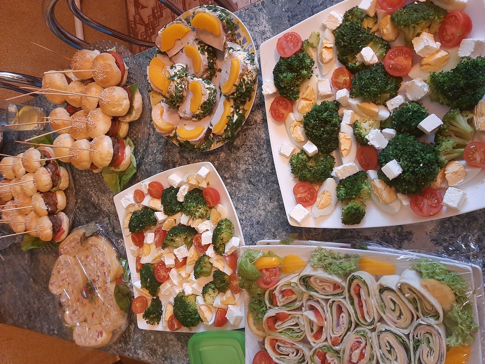 Różnorodne przekąski na talerzach: mini-burgery na szpadkach, roladki z szynką i warzywami, sałatka z brokułami, pomidorami i serem feta, oraz plastry mięsa z brzoskwiniami i ziołami.