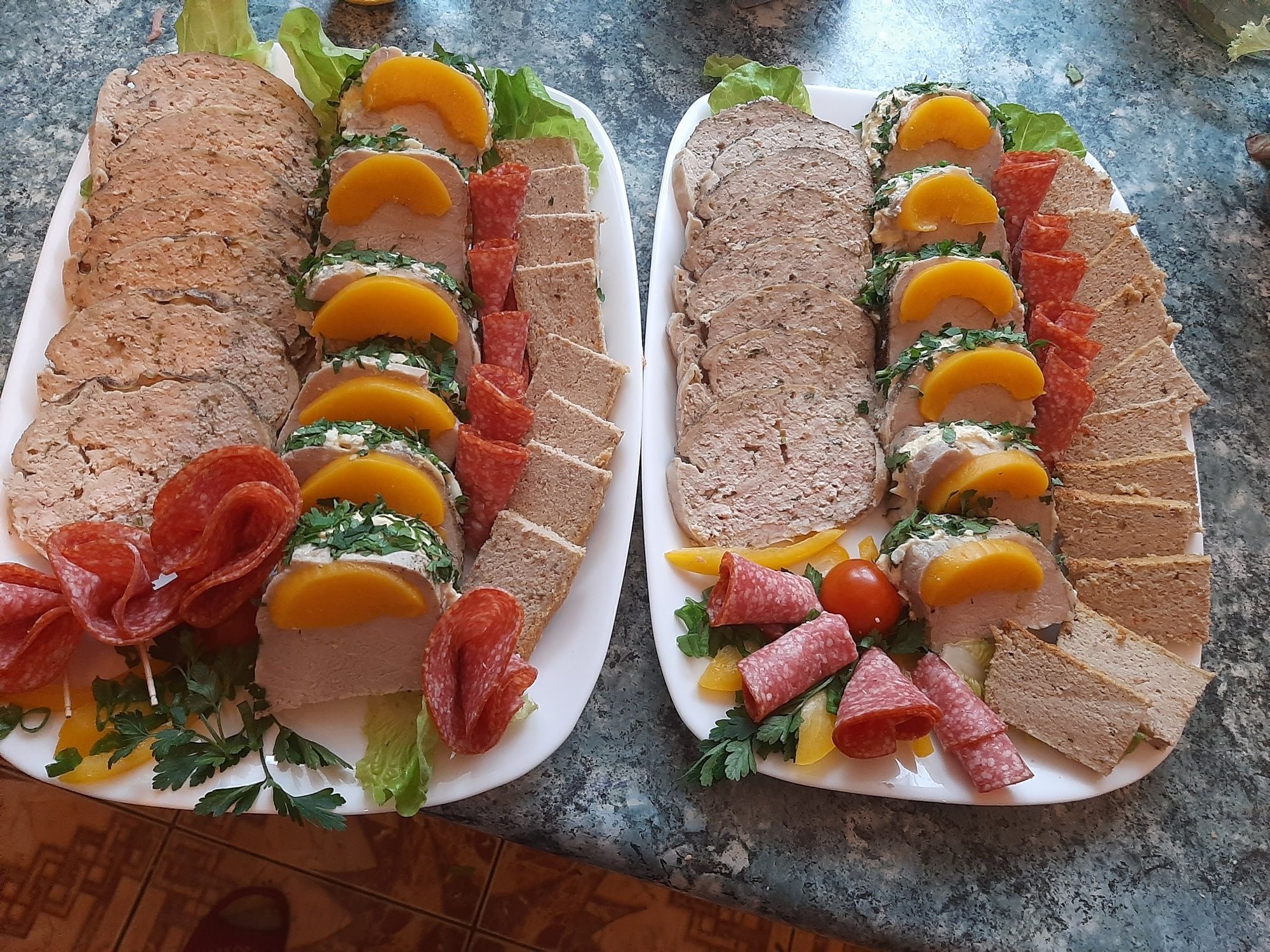 Dwa białe półmiski z pasztetem, salami, pieczywem i brzoskwiniami, udekorowane zieloną pietruszką i sałatą, na tle marmurowego blatu.