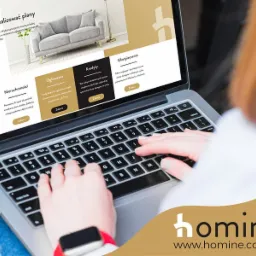 Osoba korzysta z laptopa, na ekranie strona internetowa z ofertami nieruchomości, kredytów i ubezpieczeń; widoczny fragment strony z kanapą w tle.