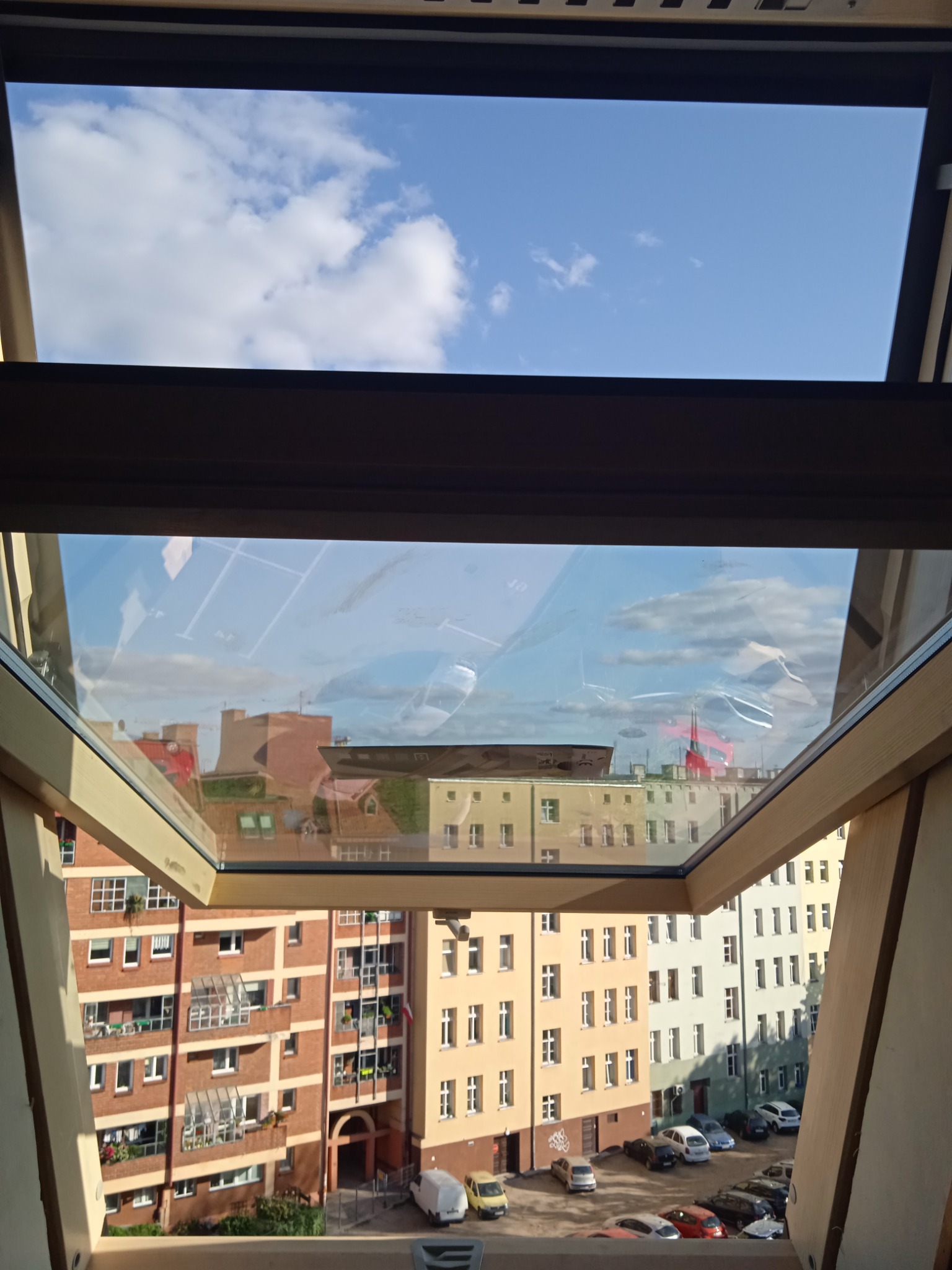 Otwarty okno dachowe z widokiem na błękitne niebo z chmurami oraz zabudowę miejską z ceglanymi i pastelowymi budynkami.