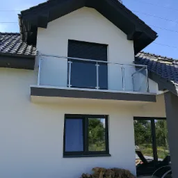 Nowoczesny dom z balkonem zabezpieczonym szklaną balustradą z metalowymi słupkami, widok z dołu na białą elewację i ciemną dachówkę.