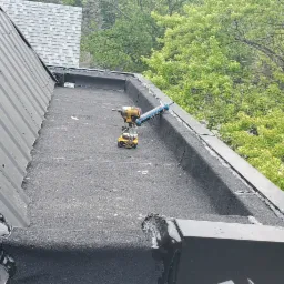 Czarny, płaski dach z papy z narzędziami: wiertarko-wkrętarka i aplikator do uszczelniacza silikonowego, widoczne ślady świeżo położonego uszczelniacza, w tle blaszany dach skośny i korony drzew.