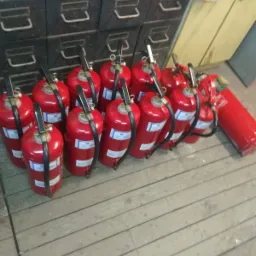 Rząd czerwonych gaśnic proszkowych stojących na drewnianej podłodze, z metalowymi szafkami w tle.