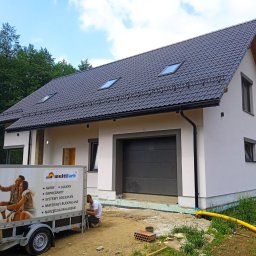 Nowbud- Usługi remontowo budowlane - Nowo wybudowany dom z ciemną dachówką i jasną elewacją, z przyczepą reklamową firmy Multifarb i pracownikiem wykonującym prace wykończeniowe przy elewacji na zewnątrz.