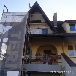 Elewacja domu w trakcie prac, widoczne rusztowania z siatką ochronną i fragment ocieplonej ściany szczytowej, balkon z donicami z wrzosami.