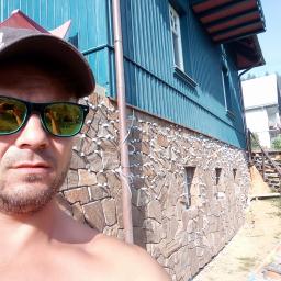Nowbud- Usługi remontowo budowlane - Mężczyzna w okularach przeciwsłonecznych robiący selfie na tle elewacji z kamienia z zamontowanymi lampkami, widoczny fragment niebieskiej drewnianej ściany budynku.