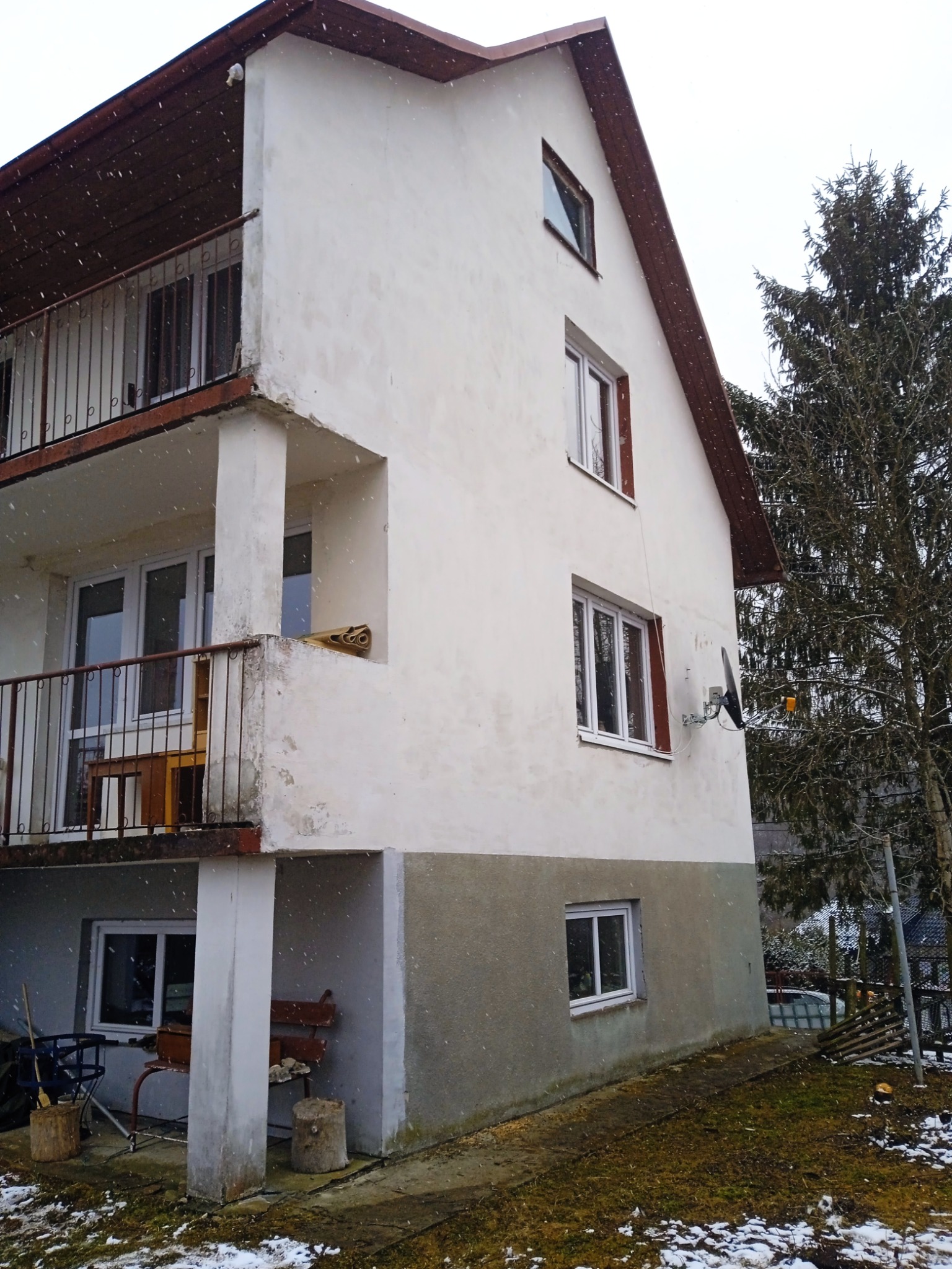 Dwupiętrowy dom z balkonami w zimowej scenerii, widoczne ślady zabrudzeń na elewacji i balkonie, antena satelitarna na ścianie, częściowo ośnieżony trawnik.