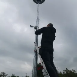 Instalator na drabinie montuje kamerę monitoringu na słupie oświetleniowym z kopułą kamery, widok z dołu na pochmurne niebo.