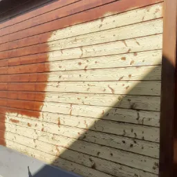 Częściowo pomalowana drewniana elewacja budynku, widoczne deski przed i po nałożeniu brązowej bejcy, z wyraźną linią podziału i widocznymi smugami.