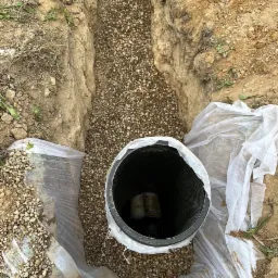 Głęboki wykop wypełniony żwirem, z widoczną rurą kanalizacyjną zabezpieczoną folią, przygotowany do dalszych prac instalacyjnych.