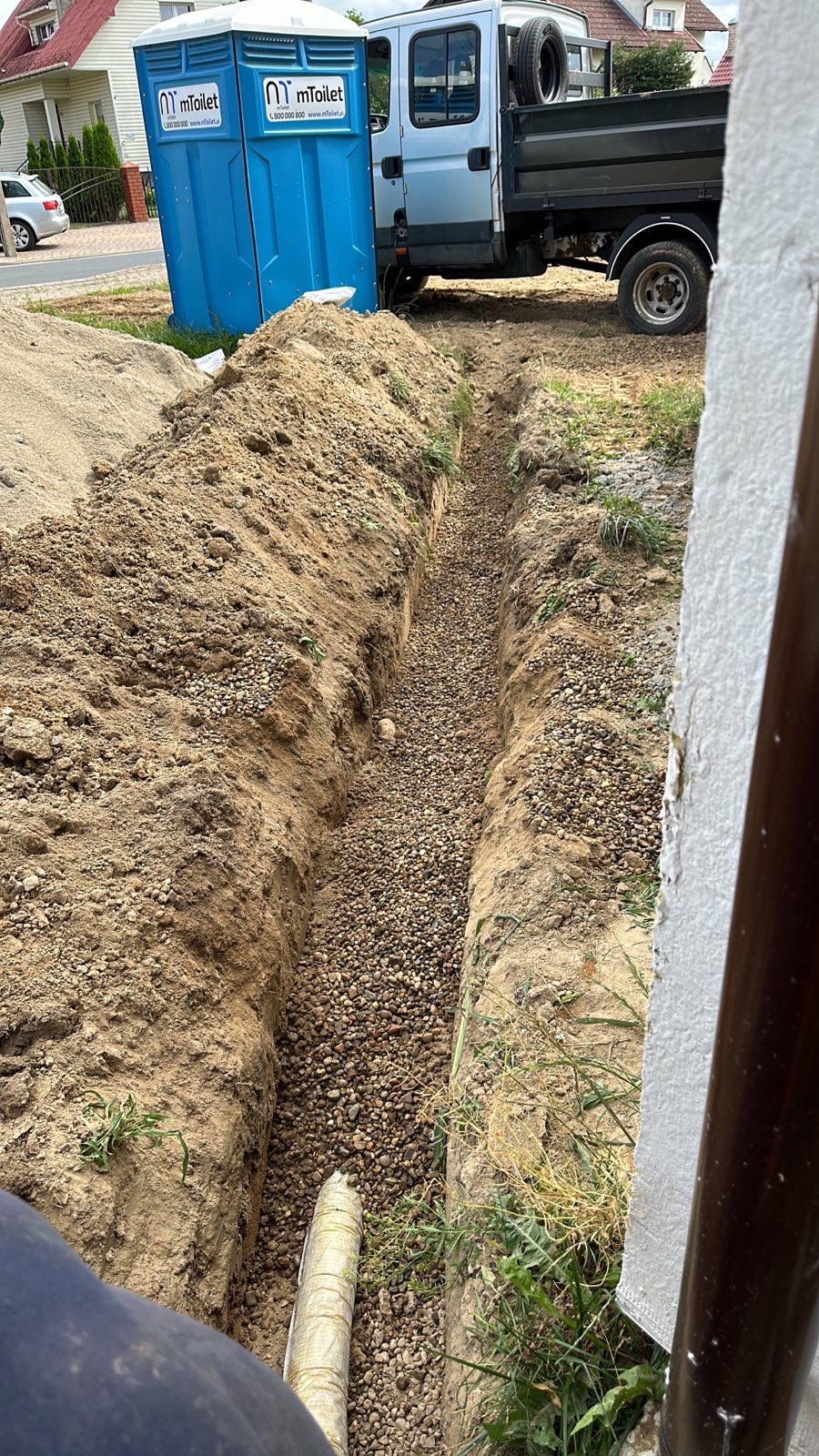 Wąski wykop wypełniony drobnymi kamieniami, widoczne dwie przenośne toalety i samochód dostawczy w tle, ziemia piaszczysta.